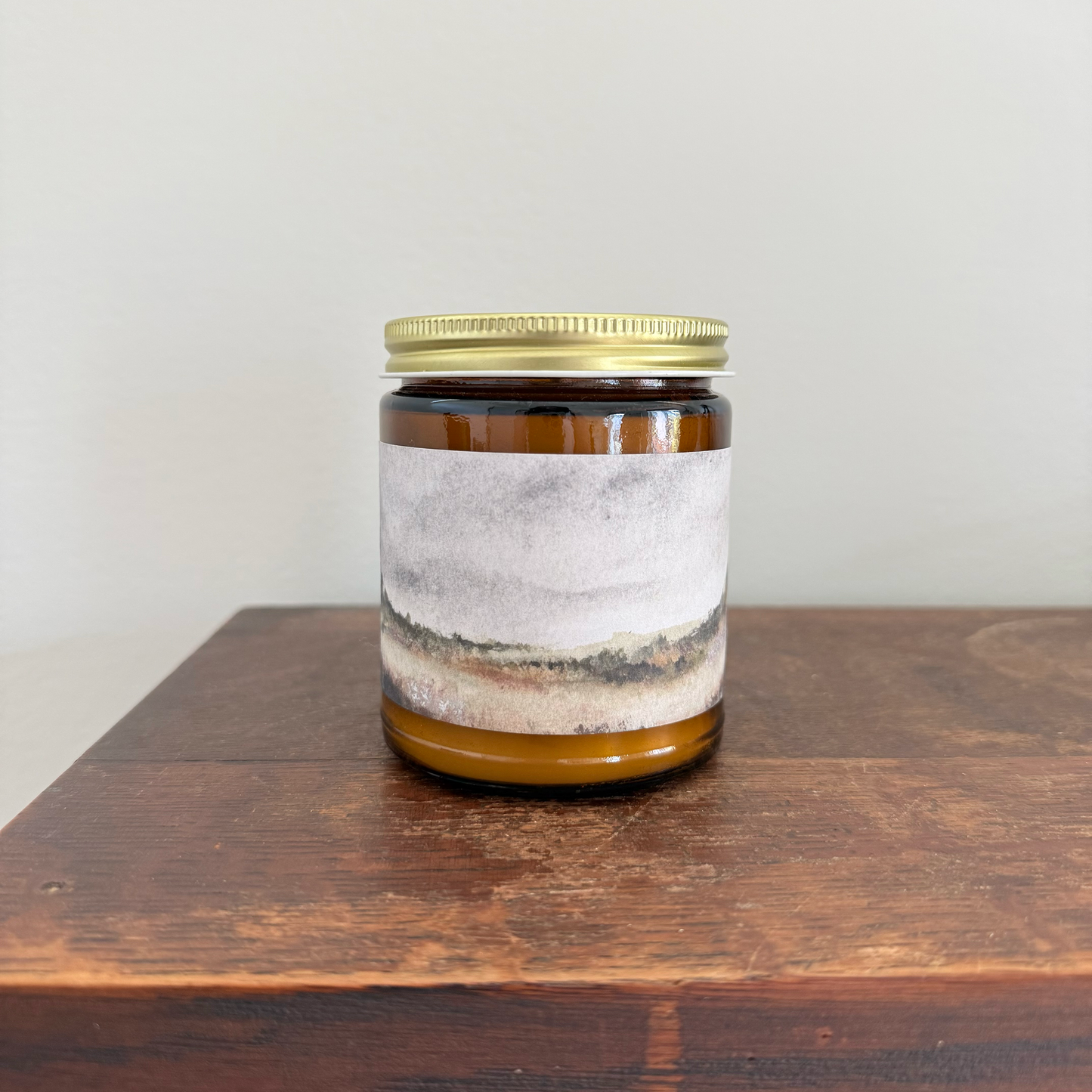 An amber glass jar candle with a gold metal screw-on lid and a textured gray and beige landscape label, sitting on a dark wooden surface against a plain light gray wall.