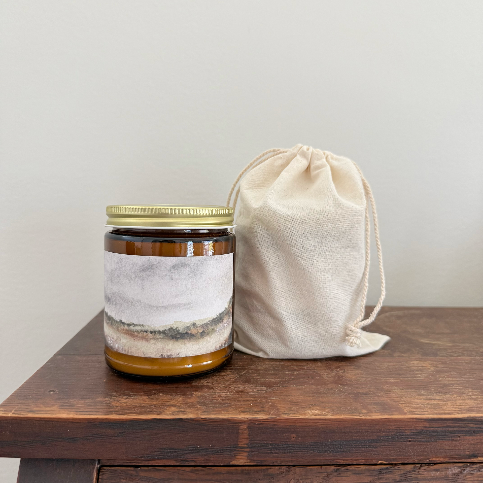 An amber glass jar candle with a gold metal lid and a scenic landscape label, standing next to a small beige cotton drawstring pouch on a dark wooden table against a light gray wall.