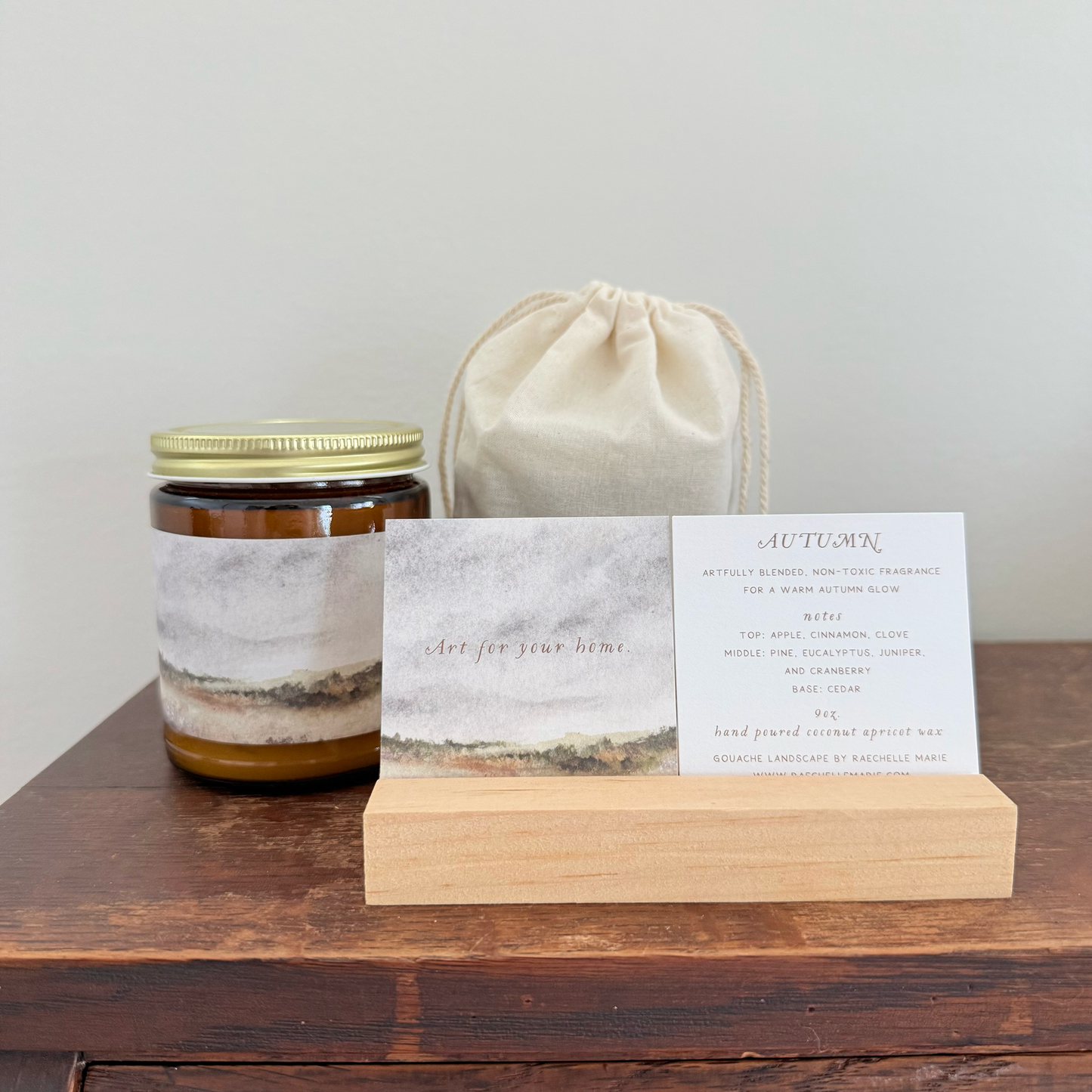 A scented candle in a glass jar with a gold metal lid and a gray-and-brown abstract label, displayed on a wooden surface next to a cream fabric drawstring bag and a printed card on a wooden stand.