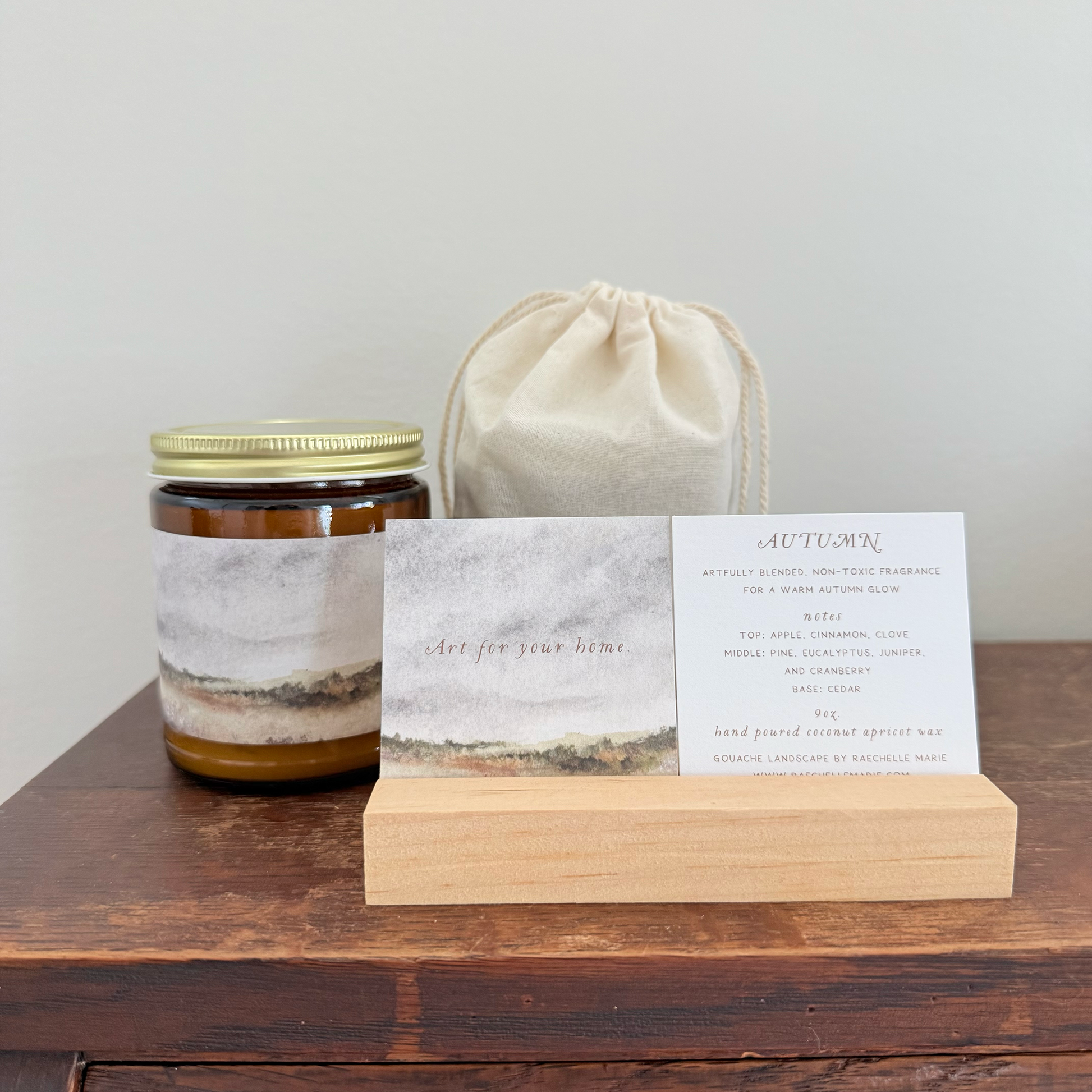 A scented candle in a glass jar with a gold metal lid and a gray-and-brown abstract label, displayed on a wooden surface next to a cream fabric drawstring bag and a printed card on a wooden stand.