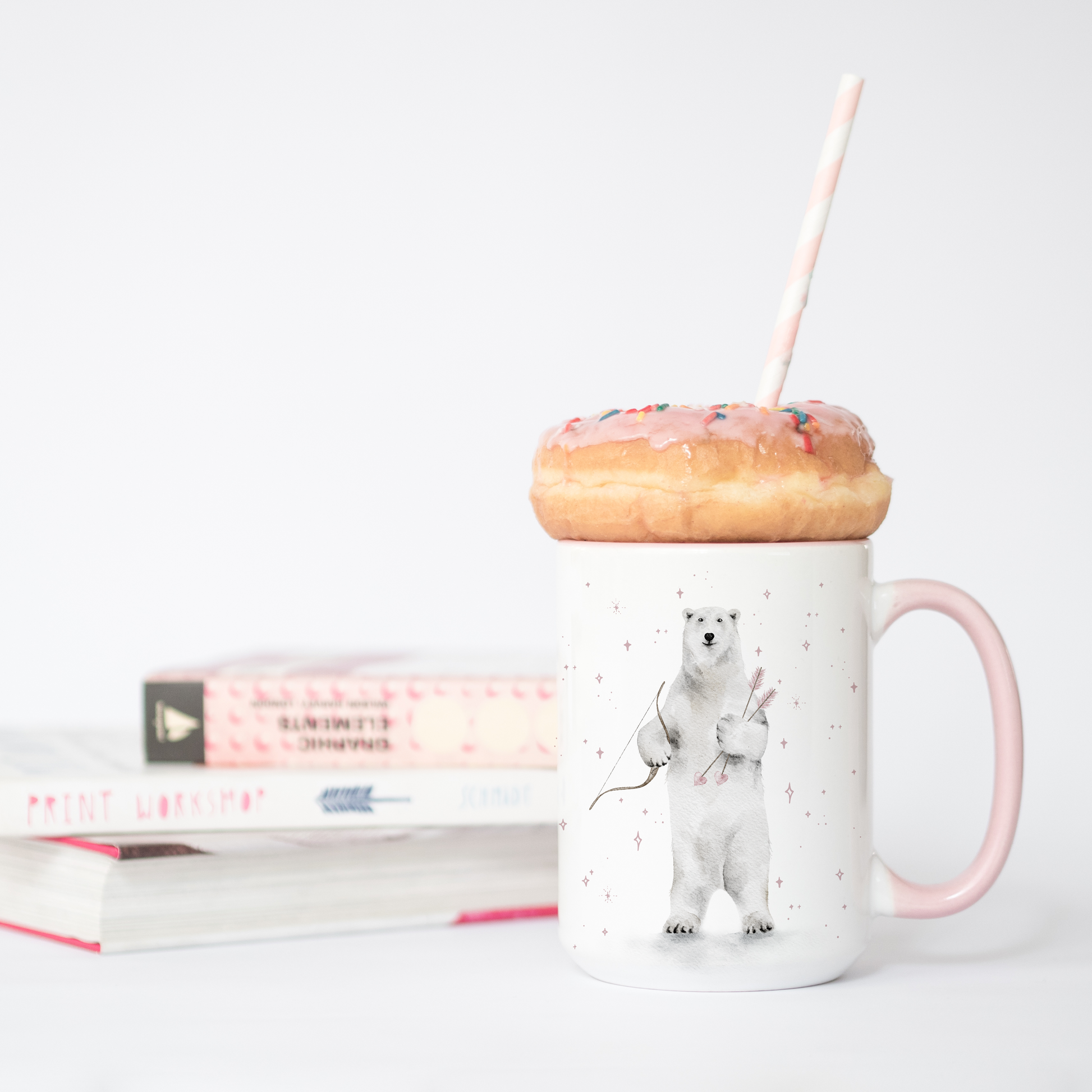 Mug with a polar bear design, pink handle, and donut on a white surface with books in the background.